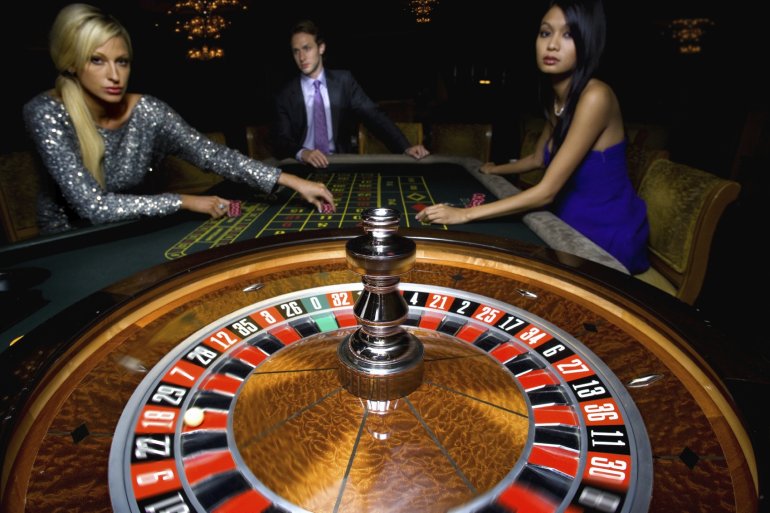 В центре фотографии колесо рулетки, а на заднем плане две красотки и мужчина - игроки казино